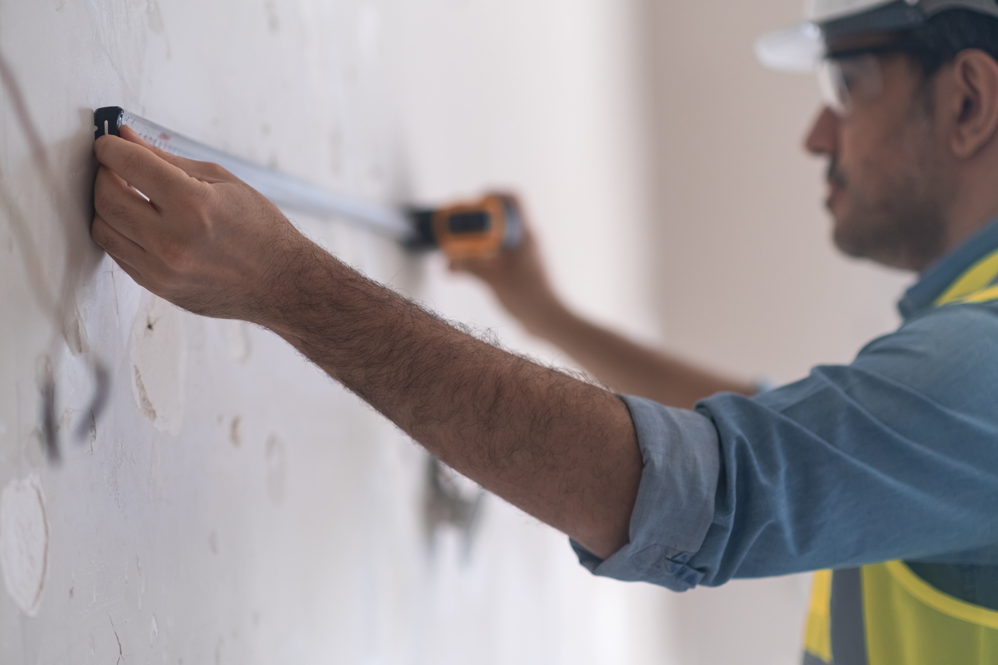 Foreman in protective goggles using measuring reel to measure shabby wall